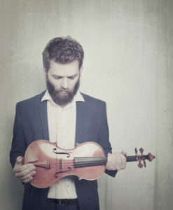 Johnny Gandelsman, a thin white man with brown hair and beard looks down at a violin which he holds in both hands at his waist.